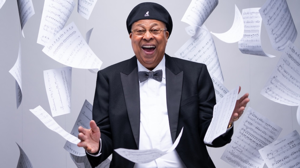 Person wearing a tuxedo and black cap smiles amidst a shower of sheet music