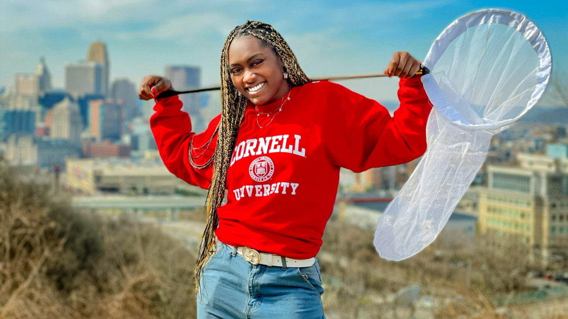 Doctoral student Sylvana Ross holds a bug net and poses in front of the Cincinnati skyline