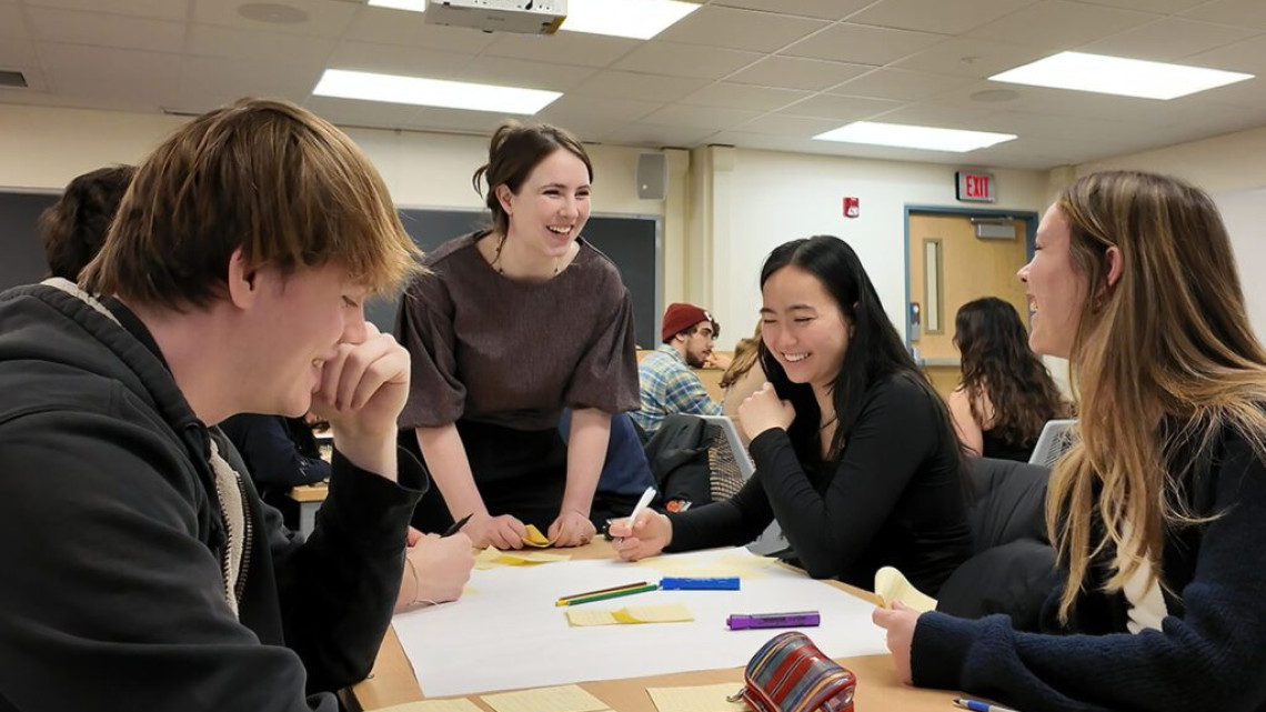 An instructor leans on a table while interacting with four students