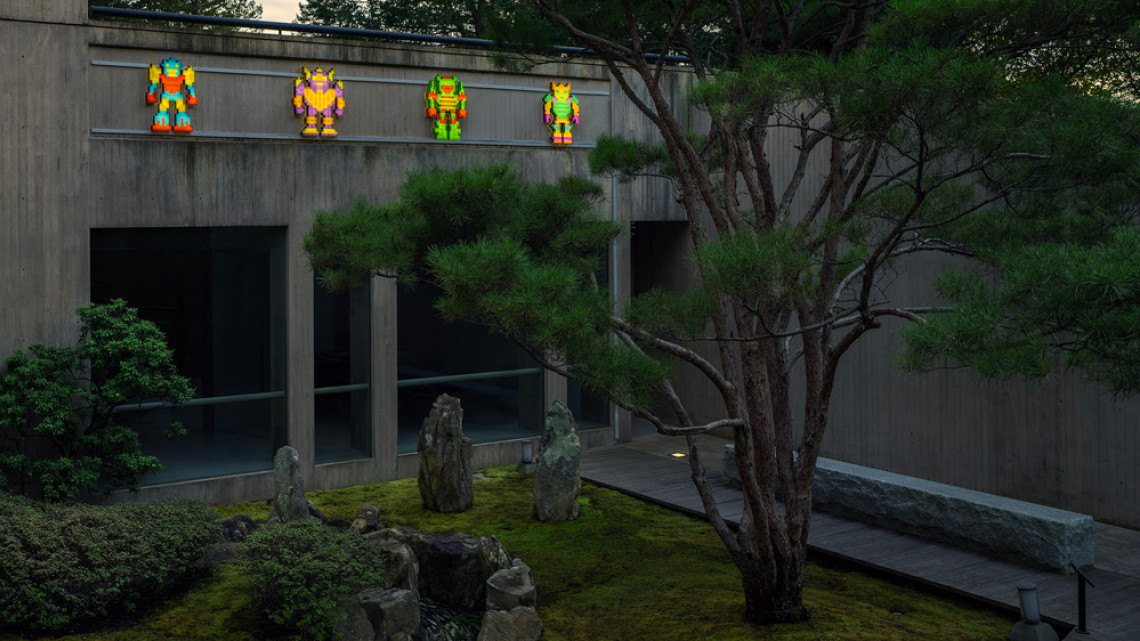 An art installation showing a Japanese garden surrounded by concrete walls with four illuminated cartoon figures mounted on a high vantage point