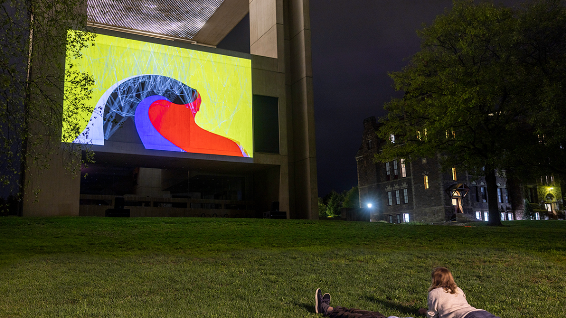 Two people laying on the grass watching a video projection on the side of a wall.