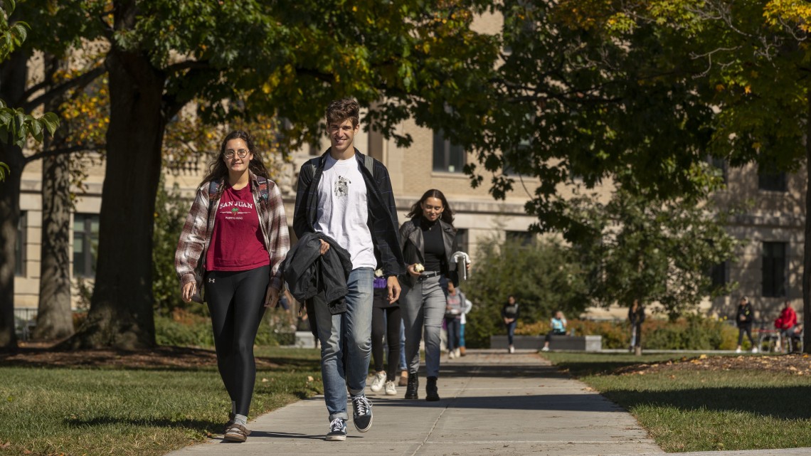 Students walking on sidewalk paths under a canopy of trees with a college building in the background.