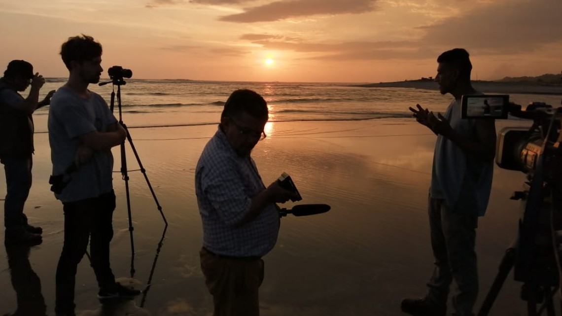 A group of people in shadowy profile film interviews while standing on a beach at sunset.