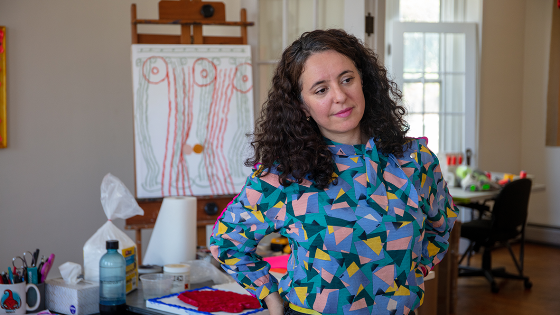 Person with long, dark, curly hair wearing geometrically patterned blouse standing with hands on hips and art supplies in background