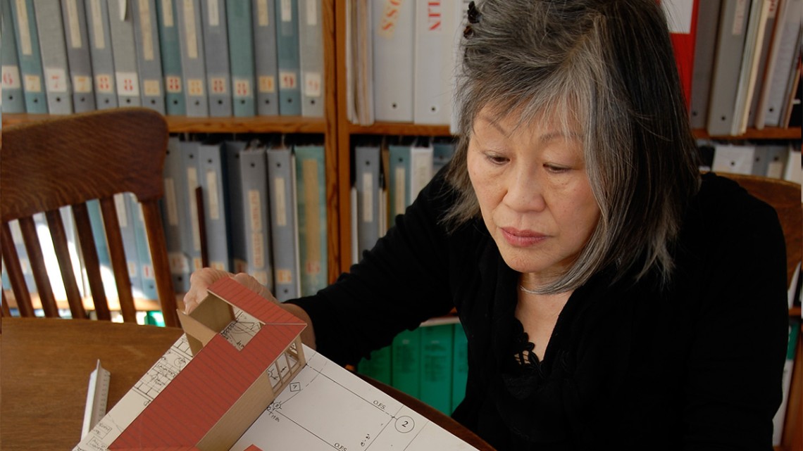 A person holding an architectural model sitting in a chair at a table with a bookshelf behind them.