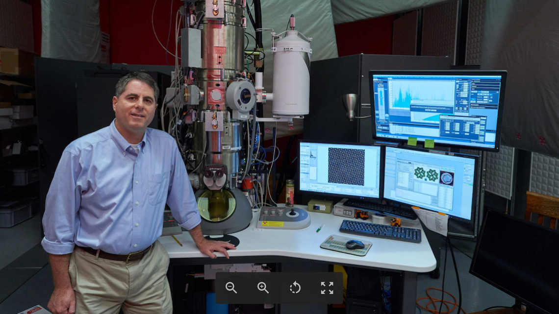 David Muller stands to the left of an electron microscope and computer monitors
