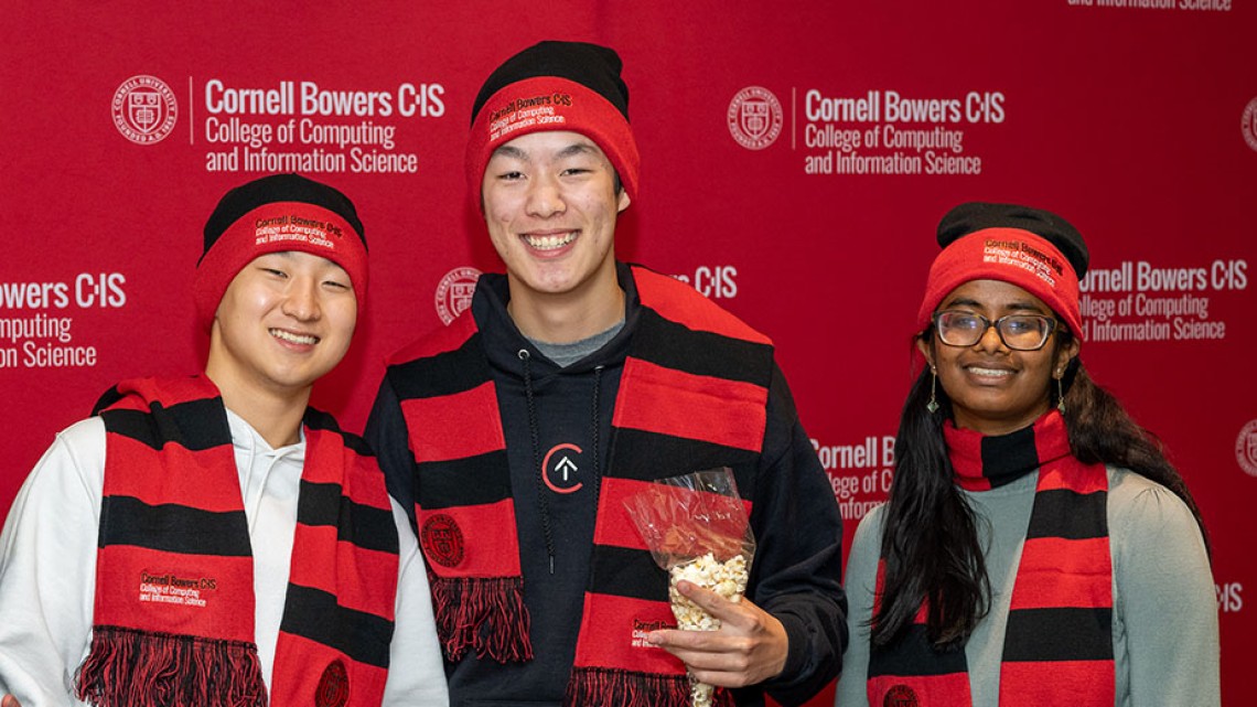 Four students in Cornell hats and scarves stand in front of a red Cornell Bowers CIS banner