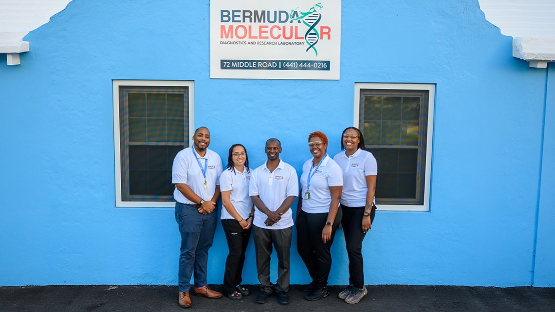 Pradeep Ambrose and Carika Weldon and 3 other people (2 women, 1 man) standing and smiling in front of a blue building with a lab sign above, "Bermuda Molecular"