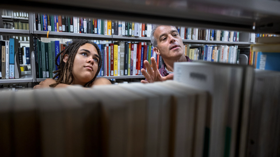 A librarian and a student look at books on a library shelf.