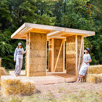 Densely packed straw bales within the interior walls of “BaleWorks,” a design-build installation by Marta Wisniewska constructed recently for BuildFest 2026 in Bethel, New York. As it evolves from a campground stage and shelter to a restroom, weathered straw will be replaced with fresh bales and repurposed for soil remediation.
