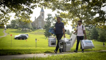 Two men carrying bedding on West Campus with McGraw Tower in the background. 