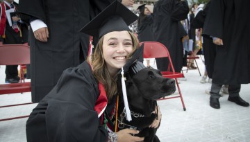 A graduate poses with a dog, both wearing graduation caps.