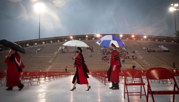 PhD candidates cross the field holding umbrellas. 