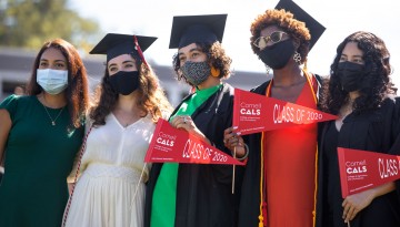 Five women wearing masks posing in cap and gown and holding CALS pennants. 