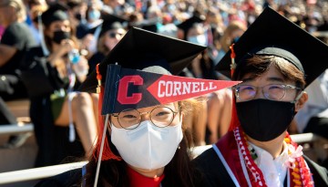Two graduates pose for a picture, one holding a small Cornell pennant. 