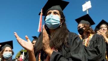 Graduates wearing masks and regalia applaud. 