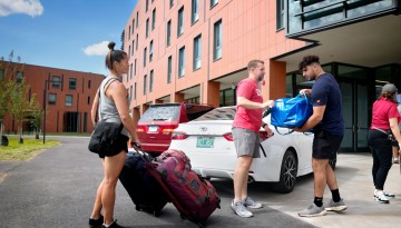 Three people carrying items and pulling luggage outside a residence hall. 