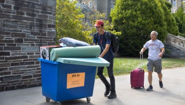 A student pushes a moving bin while his father follows behind rollin a suitcase. 