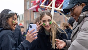 A student applies face paint as a friend holds up her phone screen. 