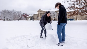 Two people build a snowman on the Arts Quad.
