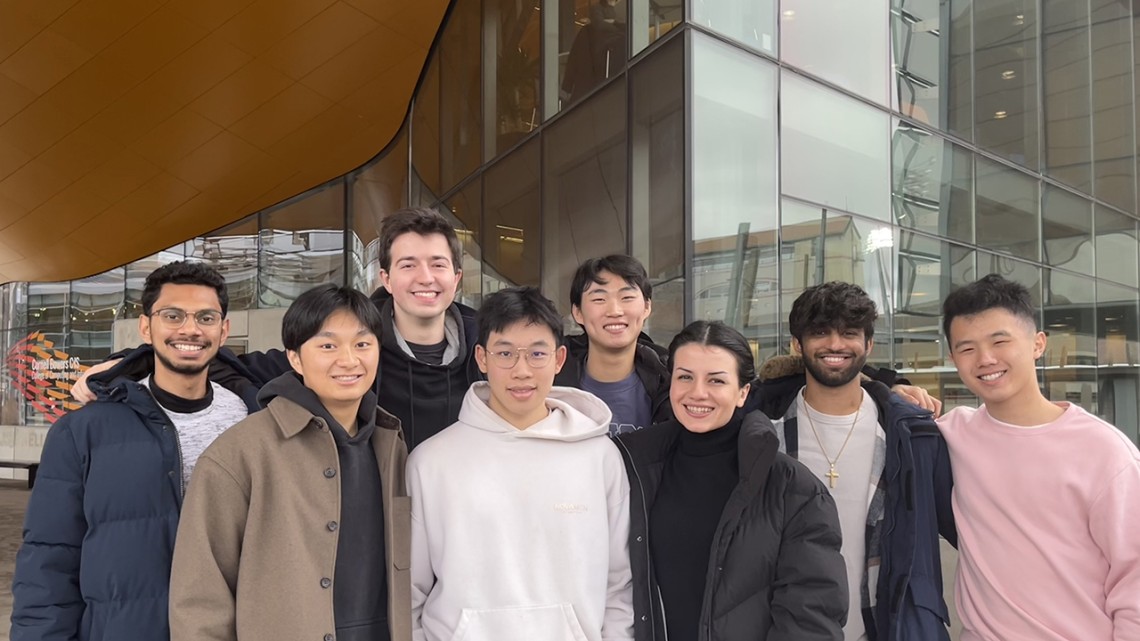 Eight students in coats stand in front of an academic building